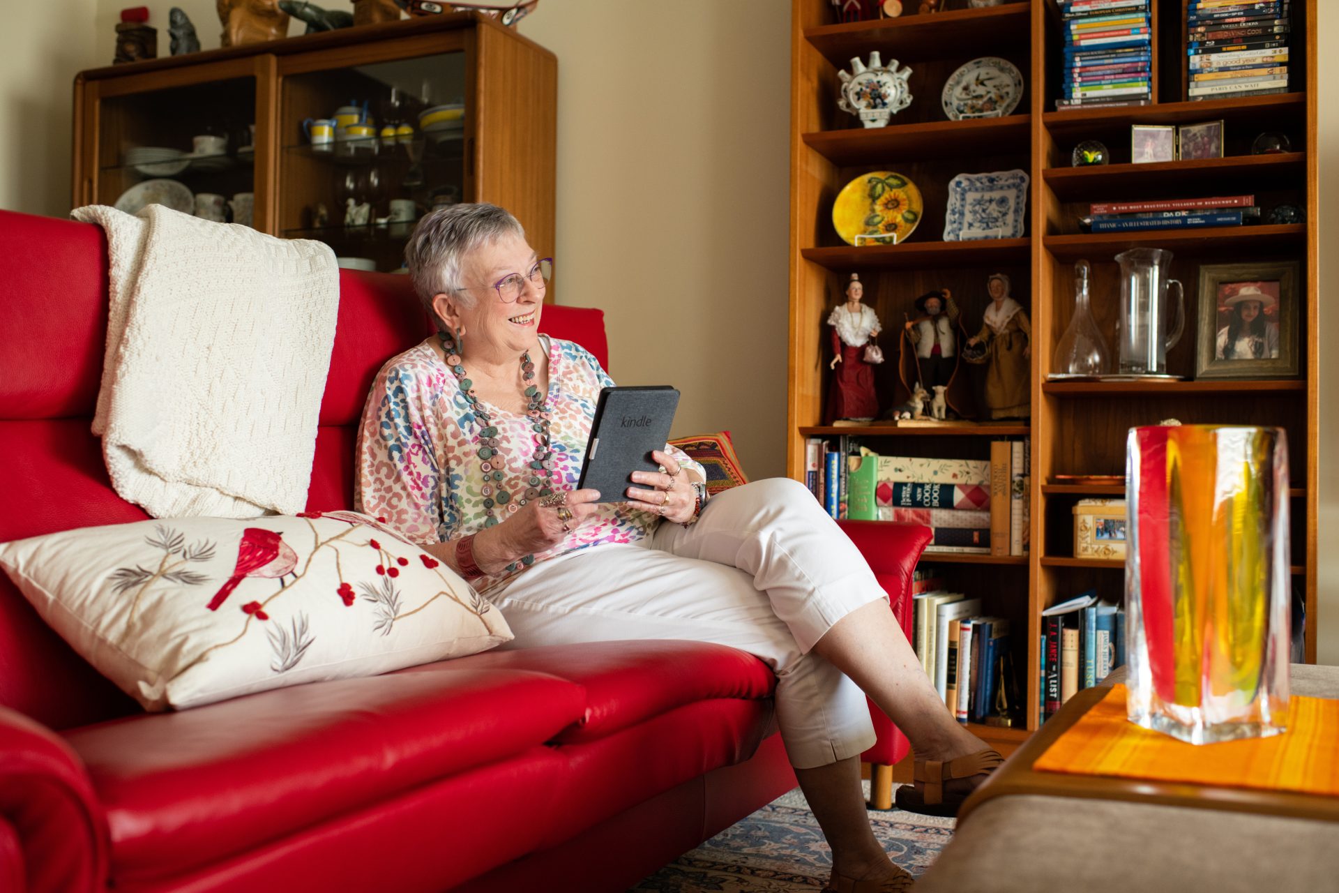 An older woman sitting on the couch reading a kindle and looking out window smiling