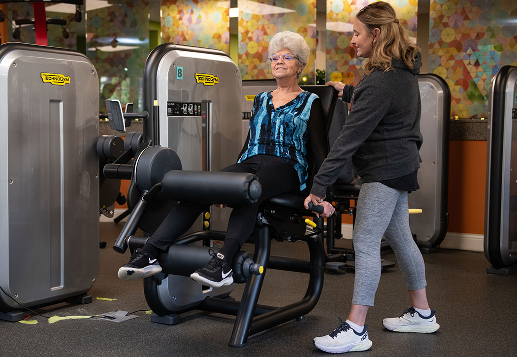 Retired women in the gym working out on leg machine with a trainer with her